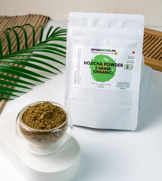 A package of Hojicha Powder labeled as organic, with a small open container of the powder visible in front, placed on a white surface with a green leafy decoration to the side.