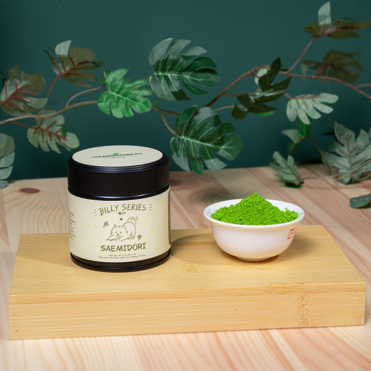 A jar labeled 'BILLY SAEMIDORI' sits on a wooden block next to a small white bowl filled with bright green powder, set against a dark green background with decorative green leaves.
